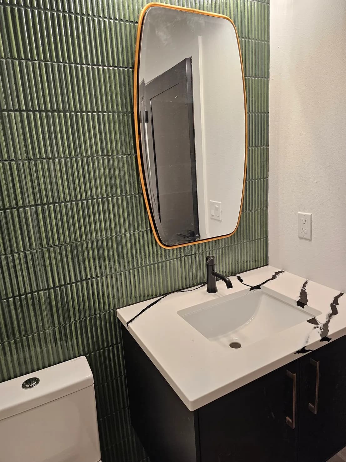 Half bath with green vertical fluted tile accent wall, gold-rimmed asymmetric mirror, black vanity with marble top