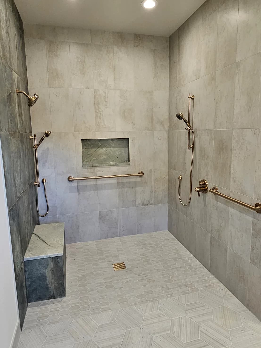 Large accessible walk-in shower with natural stone-look tile, herringbone floor, brass grab bars, corner bench, and recessed niche