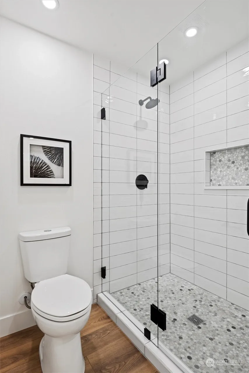 Corner shower with white stacked subway tile, pebble mosaic floor, frameless glass door, black fixtures, and penny tile accent niche