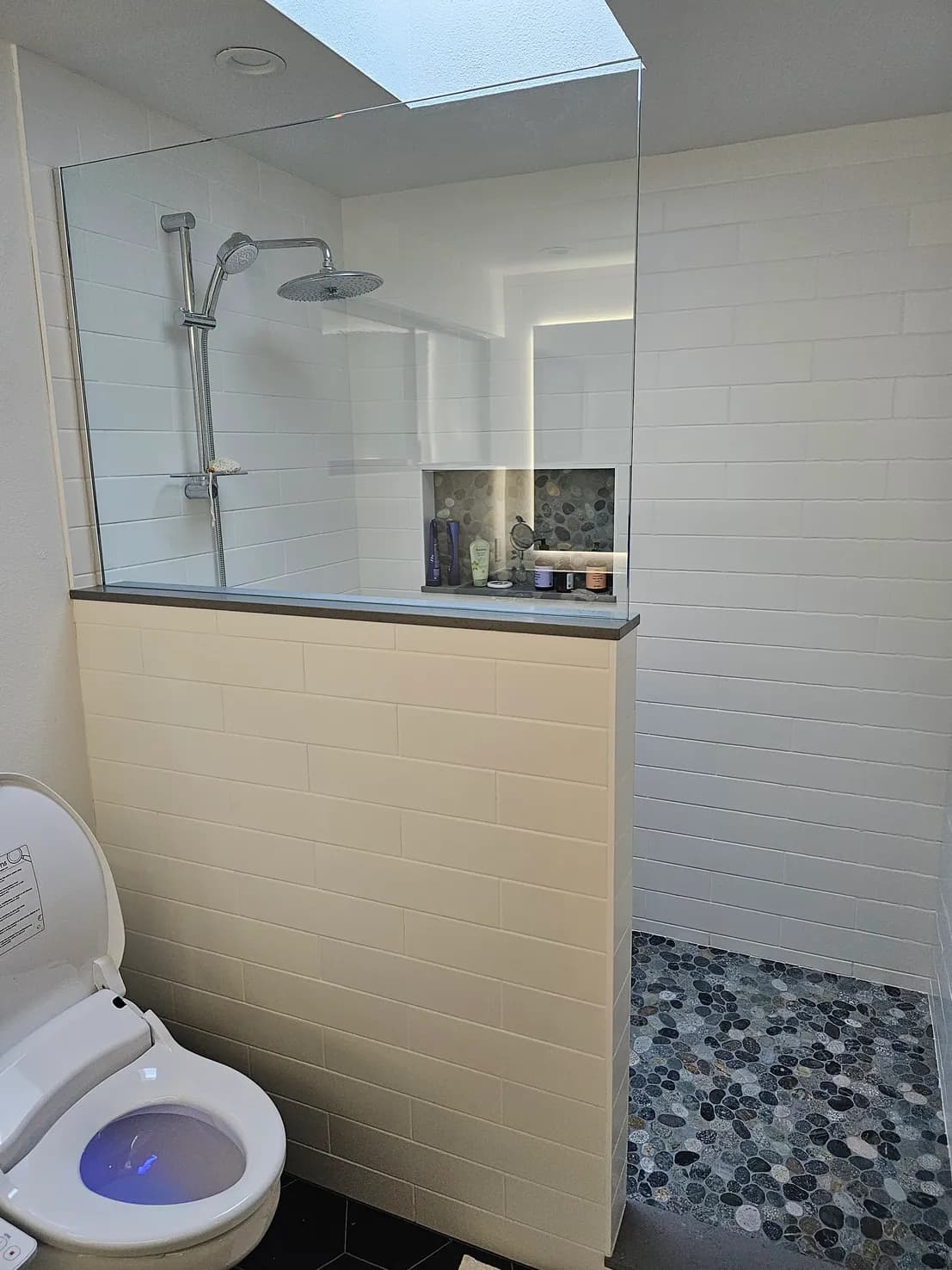 Compact bathroom with half-wall shower partition, stacked subway tile, pebble floor, rain shower head, and smart toilet