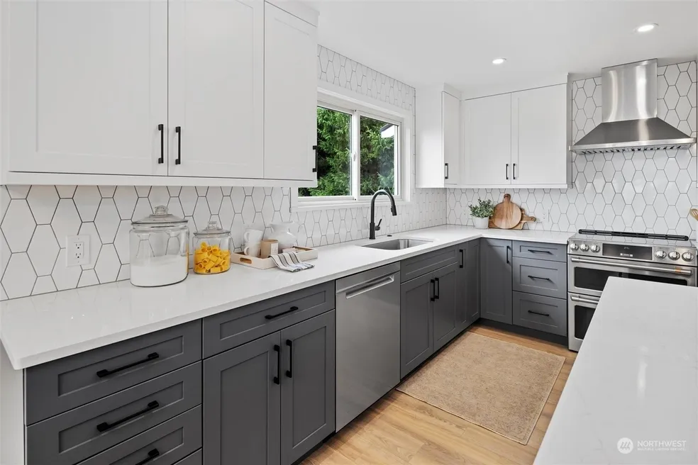 Kitchen perimeter with white upper cabinets, dark lower cabinets, hexagonal backsplash, window over sink, and stainless range
