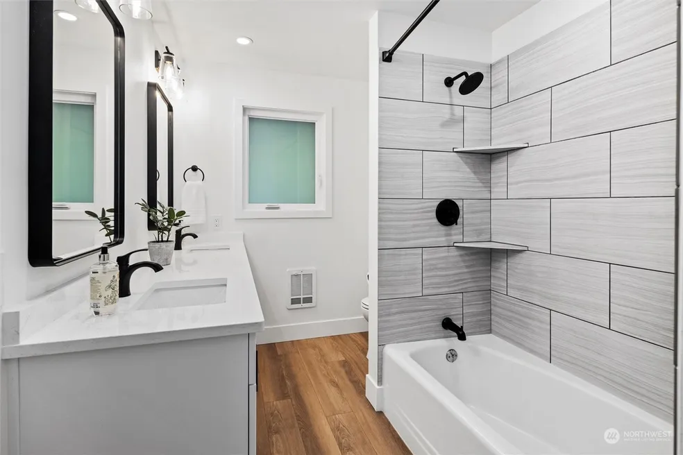 Hall bathroom with grey wood-look tile tub surround, corner shelves, matte black fixtures, and light grey vanity